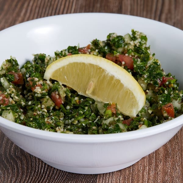 A white bowl of ShouFi MahFi Mediterranean tabbouleh salad with a lemon wedge on a wooden table.